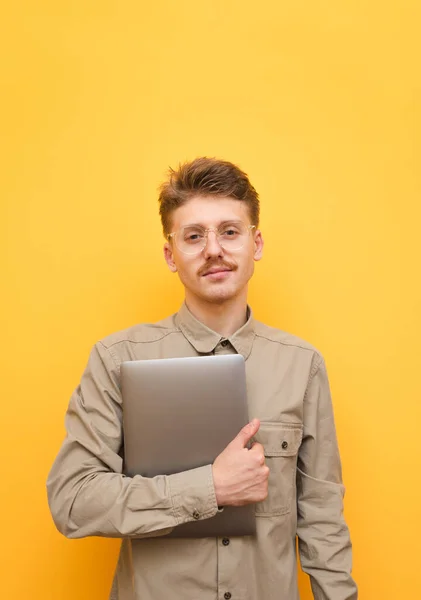 Портрет молодого программиста в футболке с ноутбуком на желтом фоне, в рубашке и очках, смотрящего в камеру и мило улыбающегося. Человек с усами держит ноутбук в руке и позирует — стоковое фото