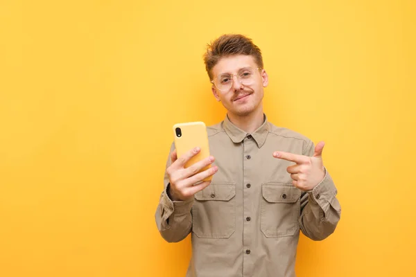 Χαρούμενος μαθητής με γυαλιά και πουκάμισο στέκεται σε κίτρινο φόντο με ένα smartphone στο χέρι του και δείχνει το δείκτη του, κοιτάζει την κάμερα και χαμογελάει. Nerd χρησιμοποιεί ένα smartphone και συνιστά. — Φωτογραφία Αρχείου
