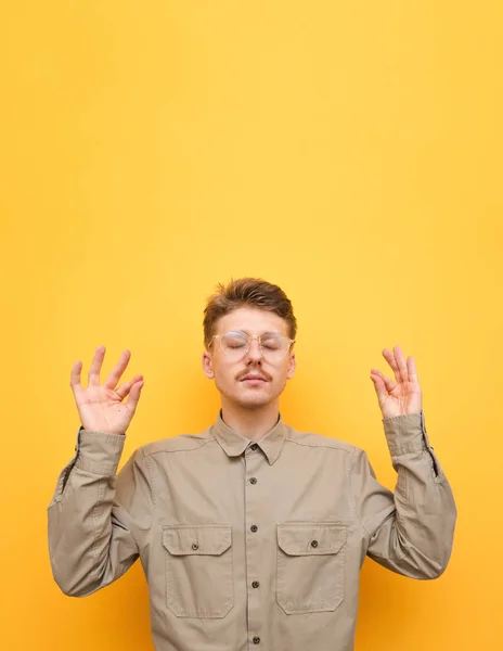 Fotografía vertical.Nerd practica yoga, aislado. Joven chico serio en camisa y gafas medita con los ojos cerrados sobre fondo amarillo . — Foto de Stock