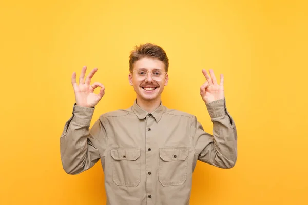 Счастливый молодой человек в рубашке и очках улыбается, смотрит на камеру и показывает большим пальцем вверх жест ОК на желтом фоне. Портрет позитивного ботаника с усами, хорошо показанным жестом, изолированным . — стоковое фото