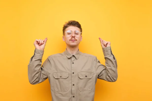 Retrato de um nerd de óculos e bigode, vestindo uma camisa, de pé sobre um fundo amarelo e meditando com os olhos fechados e rosto calmo. Jovem estudante do sexo masculino envolvido em meditação, conceito de ioga — Fotografia de Stock