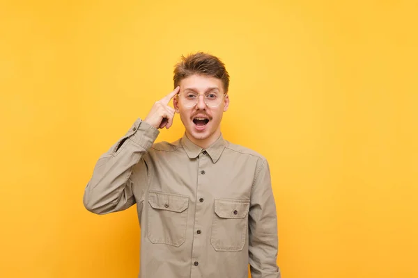 Позитивный молодой человек в рубашке и очках стоит на желтом фоне, смотрит на камеру с улыбкой и указывает пальцем на свой шаблон. — стоковое фото
