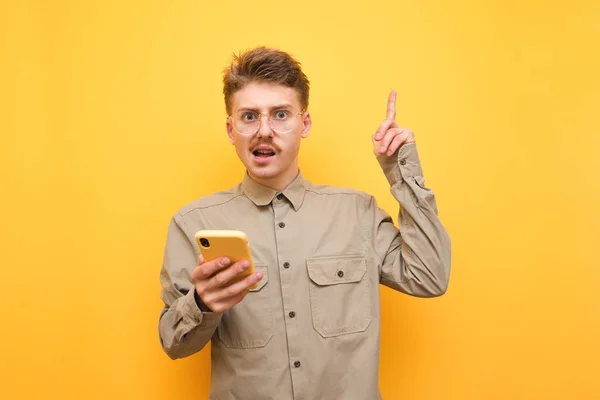 Emotional nerd in glasses and mustache stands on a yellow background with a smartphone in his hand, looks into the camera with a puzzled face and points his thumb up at copy space. Isolated — Stock Photo, Image