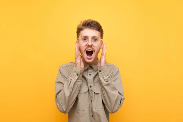 Primer plano retrato del joven sorprendido en camisa y gafas regocijándose, mirando sorprendido a la cámara con la boca abierta. Nerd alegre con bigote grita con felicidad sobre fondo amarillo. Aislado . — Foto de Stock