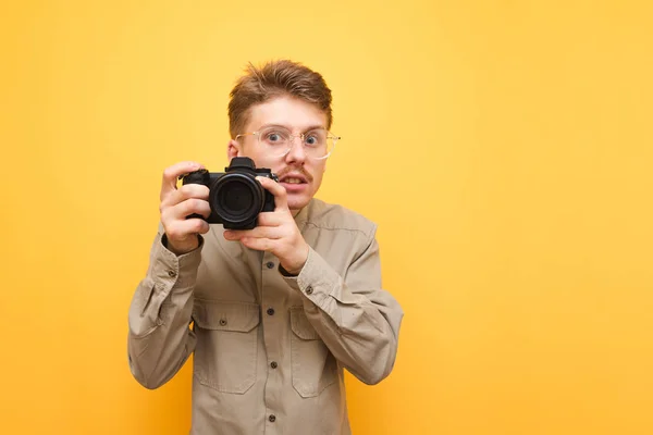 Portrait of funny stock photographer with glasses and shirt on yellow background,takes photo on camera and with serious face looks into camera.Nerd photographer with camera in hands isolated on yellow — Stock Photo, Image