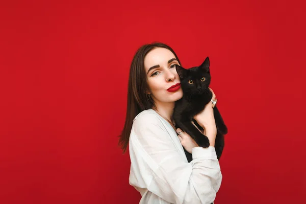 Portrait of stylish lady in casual clothes stands on a red background with a cat in her arms, posing at the camera. Beautiful woman hugging cute black cat isolated on red background. Copyspace
