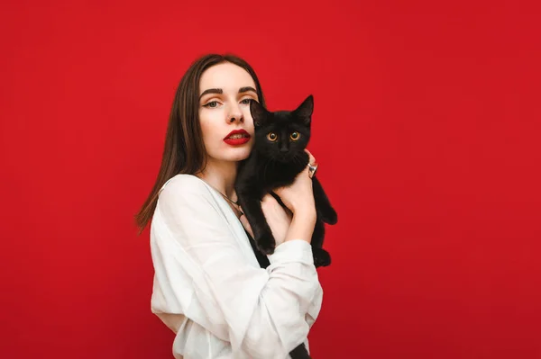 Retrato elegante de uma mulher bonita em uma camisa branca fica em um fundo vermelho com um gato preto na mão, olha para a câmera com um rosto sério. Uma rapariga a abraçar um gato, isolada. Espaço de cópia — Fotografia de Stock