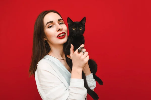 Retrato de menina feliz com gato preto em mãos no fundo vermelho, sorrindo e posando para a câmera. Bonito senhora positiva em camisa branca com animal de estimação em mãos isoladas no fundo vermelho . — Fotografia de Stock