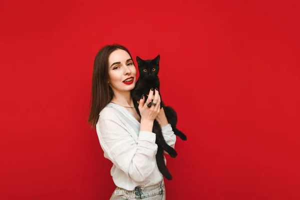 Uśmiechnięta kobieta w lekkich, luźnych ubraniach stoi na czerwonym tle z czarnym kotem w rękach, patrzy w kamerę i uśmiecha się. Szczęśliwy czarny kot właściciel odizolowany na czerwonym tle. Pojęcie zwierząt domowych. — Zdjęcie stockowe