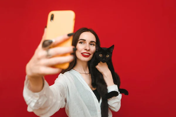 Portrait of happy woman in light clothing taking selfie with black cat on smartphone, hugging pet and looking into smartphone camera on red background. Cat owner makes a selfie with a pet. Isolated.