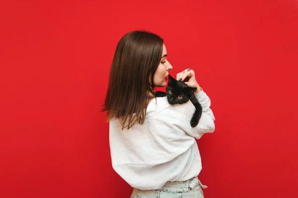 Portrait of a woman from the back holding a cat on her shoulders hugging and stroking, the cat looks into the camera. Back of beautiful girl with black cat on shoulders on red background.