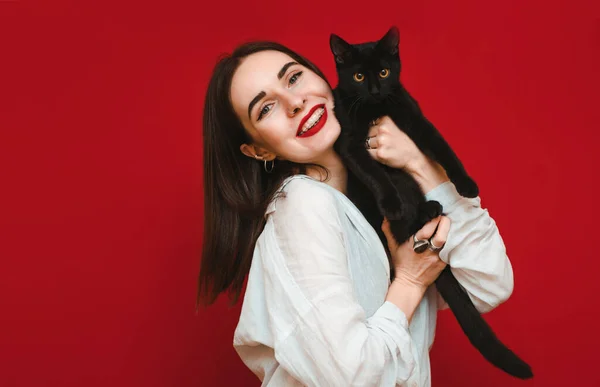 Portrait happy attractive girl in stylish casual clothes on red background, holding black cat in hands,looking in camera and smiling,wearing white blouse.Cheerful lady hugs cat and rejoices.Isolated — 스톡 사진