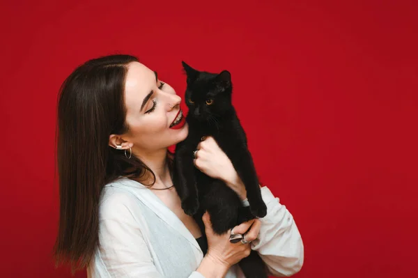 Zbliżenie zdjęcie pozytywnego właściciela z czarnym kotem na czerwonym tle, zabawy ze zwierzakiem i uśmiechem. Szczęśliwa kobieta z kotem w ramionach odizolowana na czerwonym tle. Przestrzeń chroniona — Zdjęcie stockowe