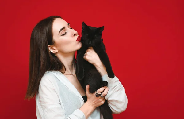 Piękna kobieta w białej koszuli przytula i całuje czarnego kota na czerwonym tle. Kobieta kocha swojego zwierzaka. Portret dziewczyny z kotem w ramionach. Odizolowane na czerwono. — Zdjęcie stockowe