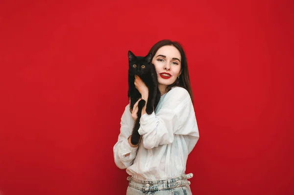 Roztomilá žena s jasným make-upem v lehkém oblečení objímá kočku na červeném pozadí, dívá se do kamery a usmívá se. Majitel a milovaný mazlíček izolován na červenou. Domácí mazlíčci. Cat and Woman — Stock fotografie