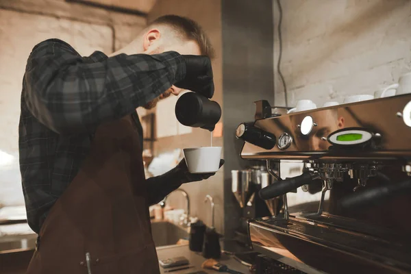 Retrato de um barista faz cappuccino em um restaurante, de pé ao lado de uma máquina de café profissional.Café proprietário prepara um café perfumado com leito.Barista derrama leite de uma xícara de pinscher com café — Fotografia de Stock