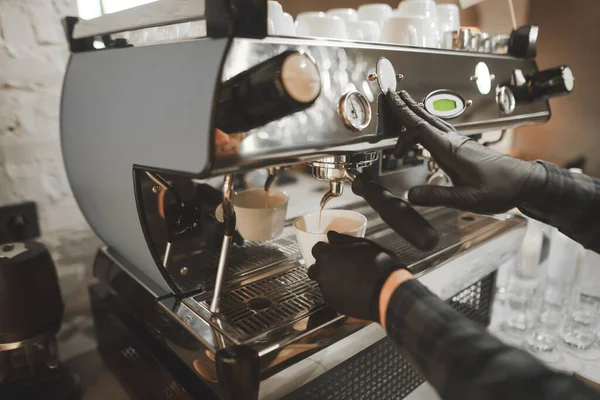 El barista profesional con guantes negros vierte café en una taza de cafetera. Cerrar foto del proceso de cocción. El café se vierte de la máquina de café en una taza. Contexto — Foto de Stock