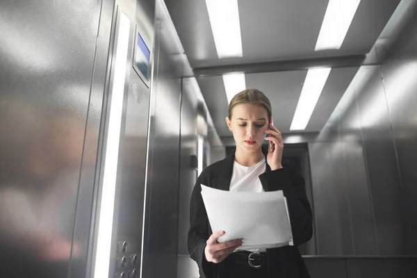 Ciddi iş kadını asansöre gazetelerde bakarak telefonla konuşuyor. Genç kadın işçiye belgeleri okuma ve smartphone Asansör tarafından konuşuyor. — Stok fotoğraf