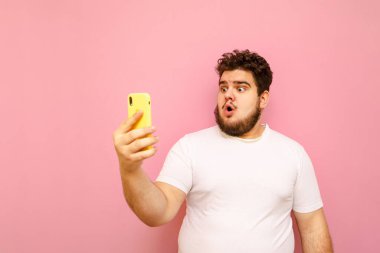 Pembe arka plandaki aşırı kilolu genç adamın portresi beyaz tişört giyen şok olmuş yüzüyle akıllı telefon ekranına bakıyor. Duygusal şişman adam selfie çekiyor, izole edilmiş..
