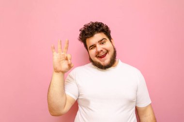 Kıvırcık saçlı ve kilolu bir adamın portresi pembe arka planda duruyor, kameraya bakıyor, gülümsüyor ve jestini gösteriyor, beyaz bir tişört giyiyor. Şişman adam iyi görünüyor.