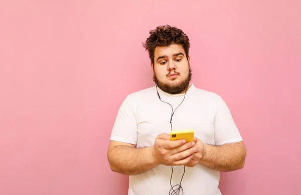 Calm curly overweight man in white t-shirt and beard using smartphone and listening to music on pink background. Fat man listens to music in a headset and holds. in the hands of a smartphone