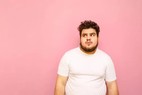 Funny fat man in white T-shirt and curly hair with serious face isolated on pink background, looks away at copy space. Funny overweight man wearing white t-shirt stands on colored background.