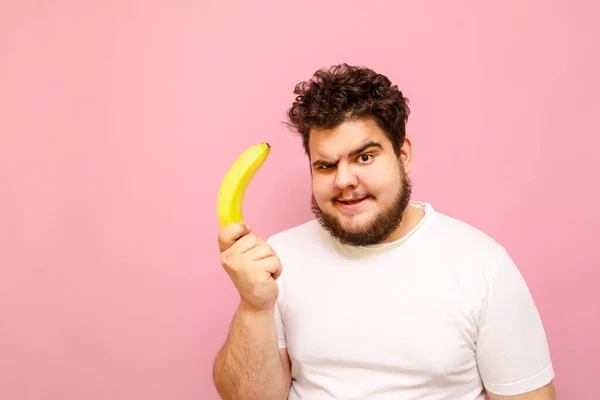 Забавный Молодой Толстяк Стоящий Бананом Руке Розовом Фоне Смотрящий Камеру — стоковое фото
