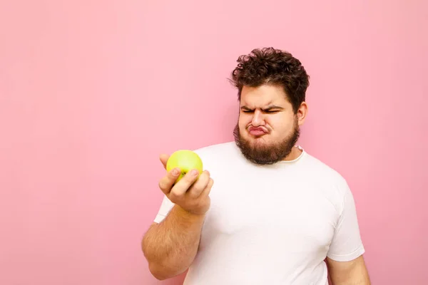 彼の手にリンゴと面白い脂肪の男の肖像画と果物を見て失望し ピンクの背景に面白い顔を作ります オーバーウェイトの男はリンゴを食べたくありません フラストレーションを示しています — ストック写真