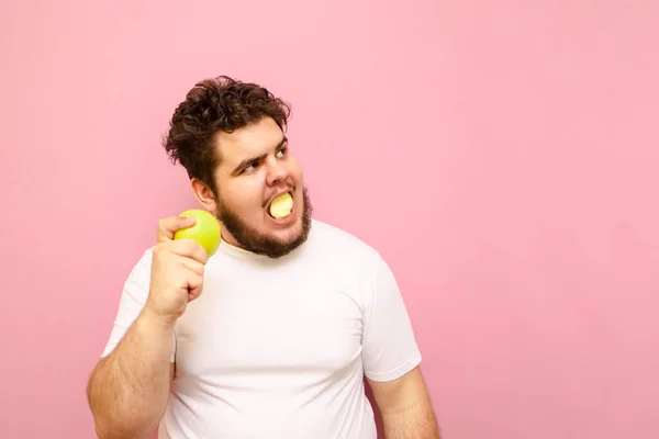 重たい顔でピンクの背景にリンゴを食べ コピースペースを離れて見る太りすぎの男 脂肪若いです男食べる緑のリンゴ上の食事療法見て驚きでザ空のスペース — ストック写真