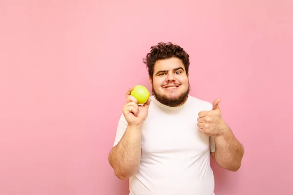 Funny bearded fat man in a white T-shirt stands with an apple in his hands on a pink background, shows his thumb up and smiles with sarcasm. Funny fat man on a diet isolated with an apple in his hand