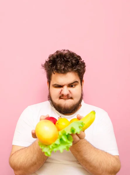 一个心怀不满的年轻胖子的画像 手里拿着水果 背景是粉色的 满脸怒火地看着生食 正在节食的胖子不想吃水果 垂直方向 — 图库照片