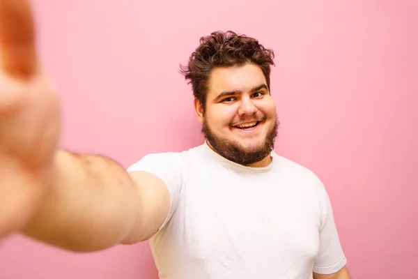 Happy overweight man taking selfie on pink background, looking into camera and smiling. Funny fat man taking his photo on camera. Isolated. Copy space