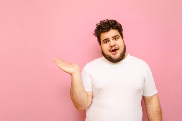 ピンクの背景に白いTシャツを着た面白い巻き毛の太った男は 空の席に手を向け 驚くべき顔をしたカメラを覗き込む カリスマ的な大男は広告スペースのために表示されます — ストック写真