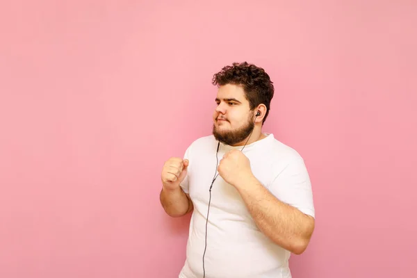 白いTシャツを着たハンサムな男と太りすぎは ヘッドフォンで音楽に耳を傾け ピンクの背景で音楽に踊り 離れて見ています — ストック写真