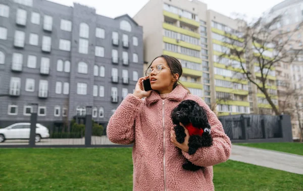 Portrait of attractive girl in pink coat walking down city street with little dog in her arms and talking on the phone. Lady walks the dog and talks on the phone.