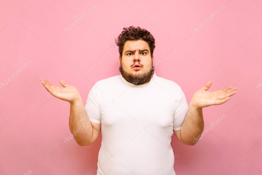 Puzzled young overweight man stands on a pink background and spreads his arms out to the sides. Confused fat guy does not know, surprise looking at the camera, isolated. Copy space