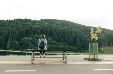 Sırt çantalı bir kız, ormanlık bir dağ manzarasının arka planında bir bankta oturuyor. Turist kızın arkası günlük kıyafetlerle arka planda dinleniyor dağlar ve kozalaklı ormanlarla