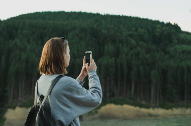 Yürüyüşçü kız günlük kıyafetlerle ve rbkzak 'la akıllı telefon kamerasıyla doğal dağ manzarası ve kozalaklı ormanın fotoğraflarını çekiyor. Yürüyüşçü kadın akıllı telefondan dağ fotoğrafı çekiyor.