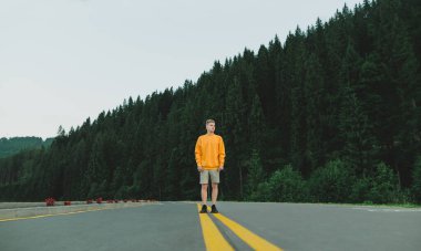 Turuncu günlük giysiler içindeki bir adamın portresi bir dağ yolunda kozalaklı bir ormanın arka planına karşı duruyor ve ciddi bir yüzle başka yere bakıyor. Turist kaldırımlı orman yolunda yürüyor..