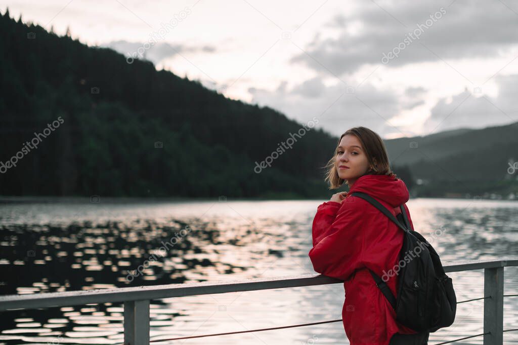 Attractive girl in a red raincoat stands by the lake and poses for looking into the camera. Hiker woman on evening walk and mountains admiring lake view.