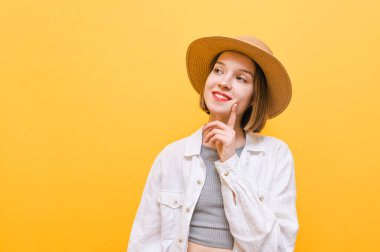 Şapkalı gülümseyen kız parmaklarıyla düşünceli bir hareket sergiliyor ve mutlu bir yüz ifadesiyle başka yere bakıyor, sarı arka planda yakın bir portre. Sarı elbiseli, düşünceli kadın..