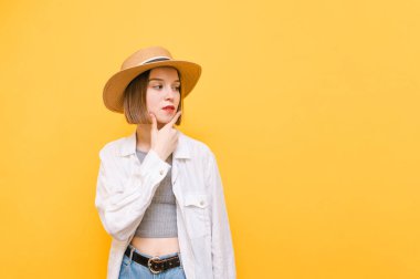 Yazlık kıyafetleri ve şapkası olan düşünceli çekici bir kız fotokopi odasına bakıyor ve düşünüyor. Düşünceli yüzlü bayan turist sarı arka planda izole edilmiş..