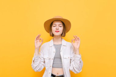Şapkalı ve açık renk elbiseli sakin bir kız elleri yukarıda, gözleri kapalı meditasyon yapıyor. Sarı arka planda yoga yapan güzel turist kız. Meditasyon.
