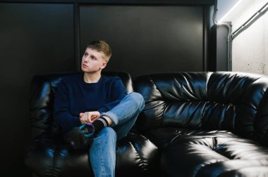 Gündelik giysiler içinde genç bir adam karanlık bir odada deri bir kanepede oturuyor, ciddi bir yüzle başka tarafa bakıyor. Uzayı kopyala Adam deri koltukta dinleniyor..