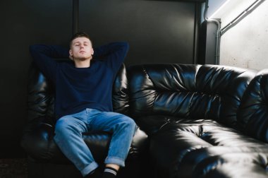 Günlük giysiler içinde yorgun bir adam karanlık bir odada deri bir kanepede oturur ve uyur. Salondaki siyah deri koltukta dinlenen genç adam. Kopya alanı