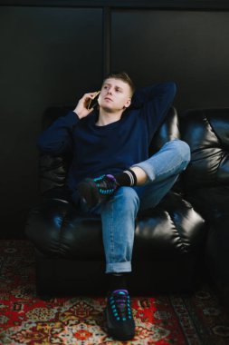Karanlık bir misafir odasında deri bir kanepede oturan ciddi bir genç adam ciddi bir yüzle telefonla konuşuyor, başka tarafa bakıyor. Dikey fotoğraf.