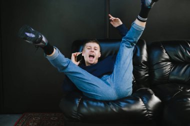 Gündelik giysiler içindeki oyuncu genç adam siyah deri bir kanepede baş aşağı yatıyor telefonda konuşuyor, kameraya bakıyor ve gülümsüyor. Adam telefonla arıyor ve kanepede eğleniyor..