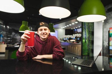 Elinde kırmızı bir bardak kola ve masada dizüstü bilgisayarla bir fast food kafede oturmuş kameraya bakıp gülümseyen gündelik kıyafetlerle gülümseyen genç adam. Bistro 'da mutlu öğrenci vakit geçirir.