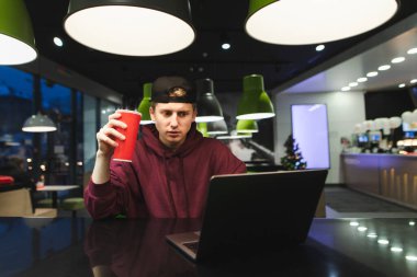 Sıradan kıyafetler giyen ciddi bir adam bir kafede dizüstü bilgisayar kullanıyor elinde soğuk bir içecek olan kırmızı bir bardak tutuyor ve bilgisayar ekranına bakıyor. Bağımsız kavram.