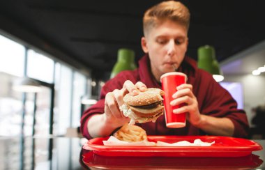 Fast food tepsisi, patates kızartması, kırmızı bardaktan içen genç bir adamın ellerinde hamburger. Fast Food Konsepti. Fast food restoranında öğrenci yemekleri..